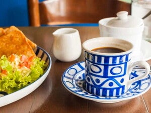 Una taza blanca y azul que contiene café y al lado un plato con ensalada verde y quesadillas