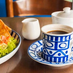 Una taza blanca y azul que contiene café y al lado un plato con ensalada verde y quesadillas