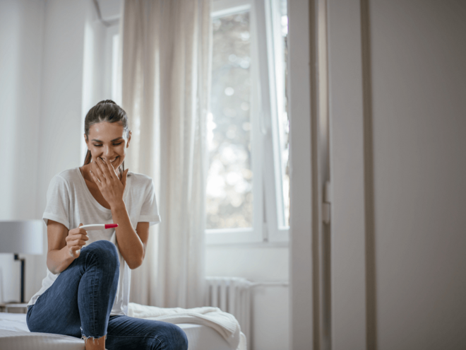 Mujer sentada en la cama sonriendo y mirando un test de embarazo